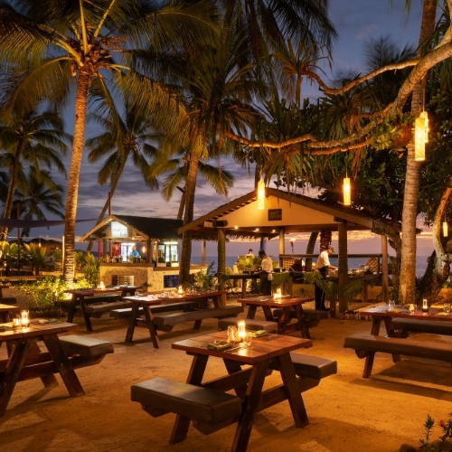 Tropical beachside restaurant at dusk, with swaying palms, glowing lanterns, wooden picnic tables, ocean views, and a warm, relaxed atmosphere.