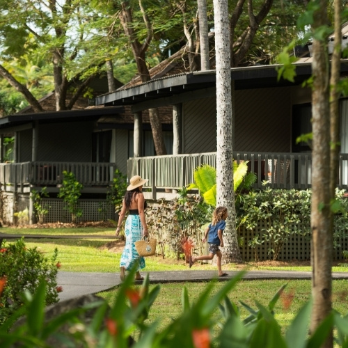 Woman in a sunhat and child walk along a tropical resort path past leafy cottages and porches, creating a relaxed vacation atmosphere.