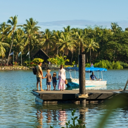 Family stands on a wooden dock beside a small boat on a tropical lake, surrounded by palm trees; bright sunlight creates a peaceful vacation mood.