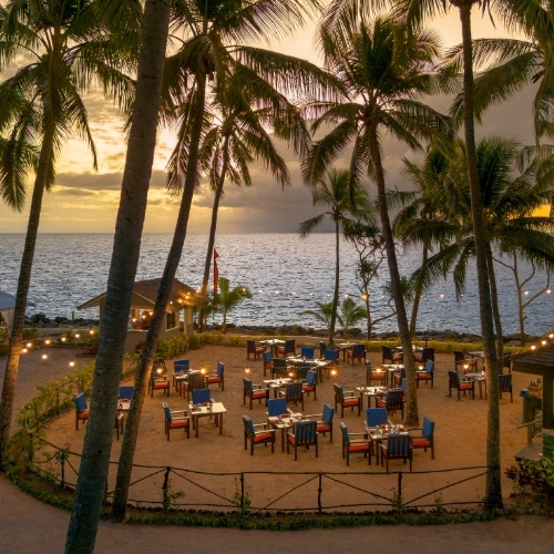 Tropical beachfront restaurant at sunset, with palm trees, ocean waves, glowing lights, and empty tables creating a warm, tranquil mood.