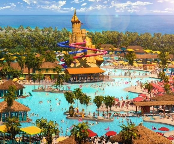 A vibrant water park featuring large swimming areas, water slides, palm trees, and sun loungers, with a clear blue sky and ocean in the background.