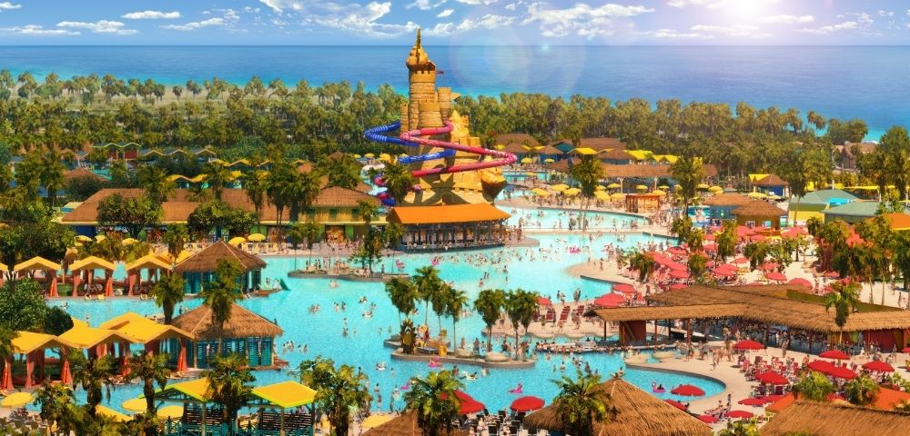 A vibrant water park featuring large swimming areas, water slides, palm trees, and sun loungers, with a clear blue sky and ocean in the background.