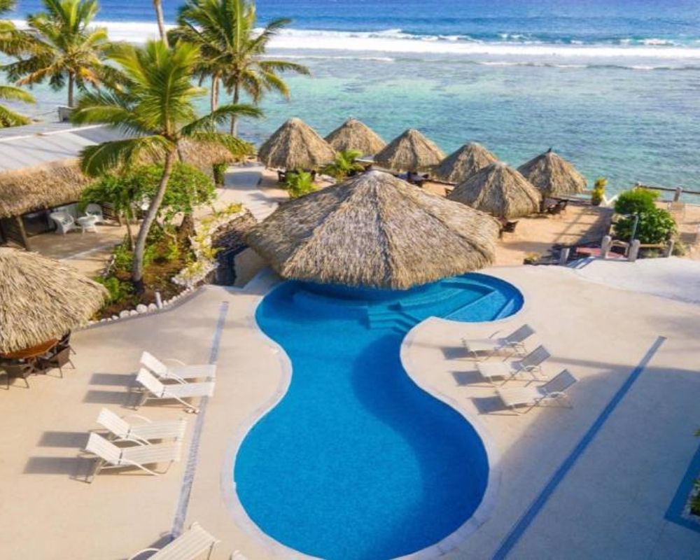 An aerial view of a tropical resort with a thatched-roof bar and pool overlooking the turquoise ocean, surrounded by palm trees and several open-air thatched-roof cabanas.