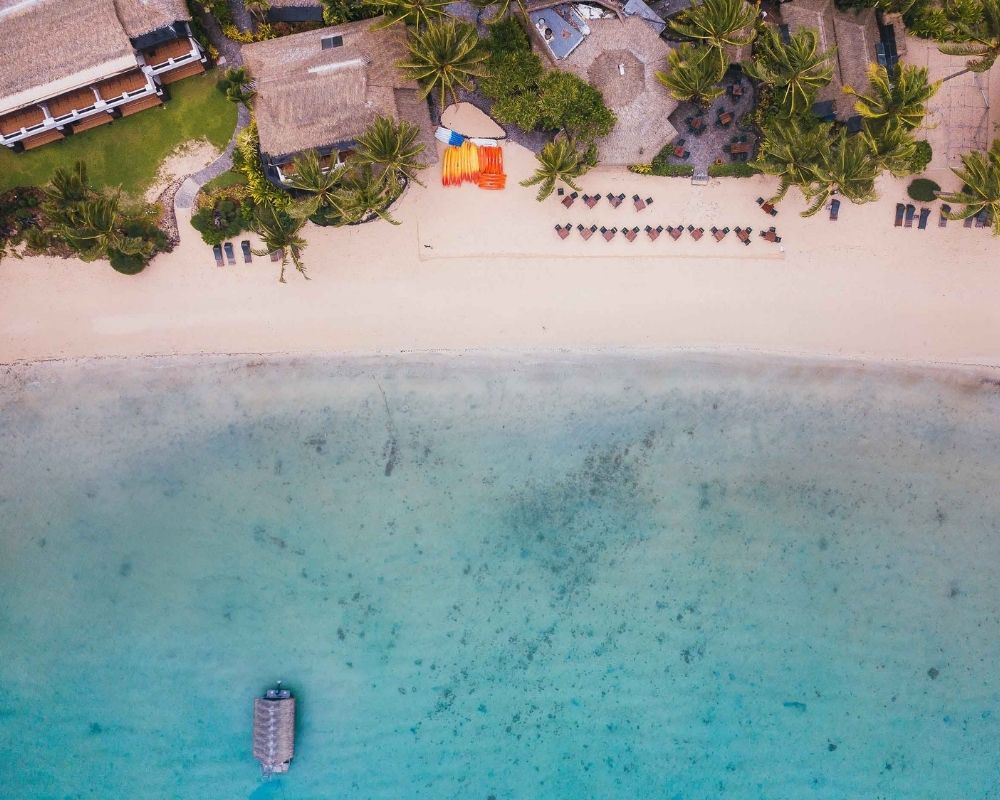 Aerial view of a tropical resort with thatched-roof bungalows, palm trees, and a sandy beach lined with lounge chairs and kayaks.