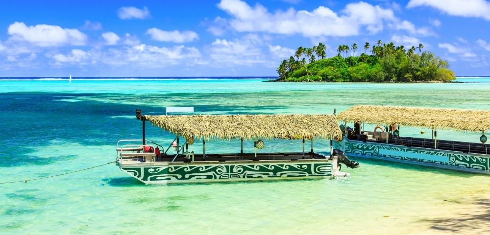 Two traditional outrigger boats with thatched roofs float in clear turquoise water, with a lush, palm-covered island in the background.