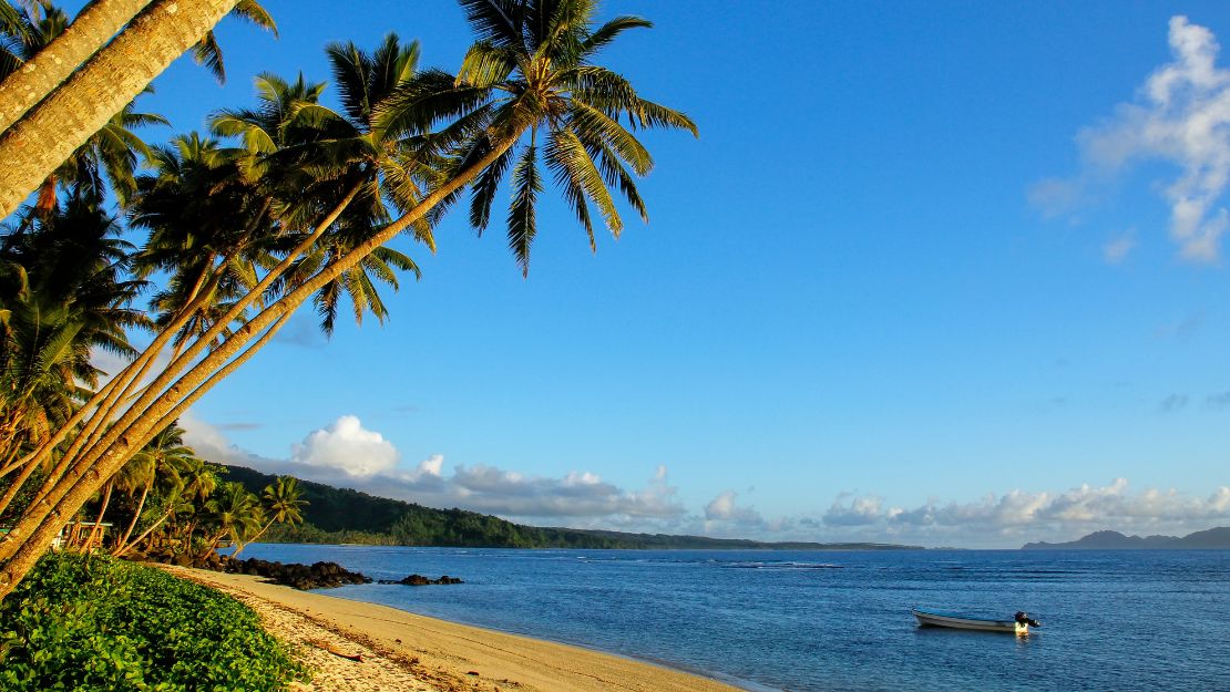 Tropical beach lined with leaning palm trees, bright blue ocean, a small boat offshore, and distant islands under a clear sky; serene mood.
