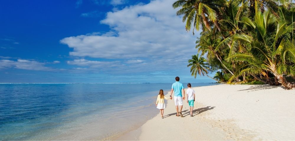 A man and two children walk along a tropical beach with palm trees and clear blue water under a partly cloudy sky.