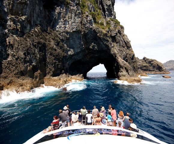 Tourists on a boat approach a large natural archway carved into a rocky cliff face over the ocean.