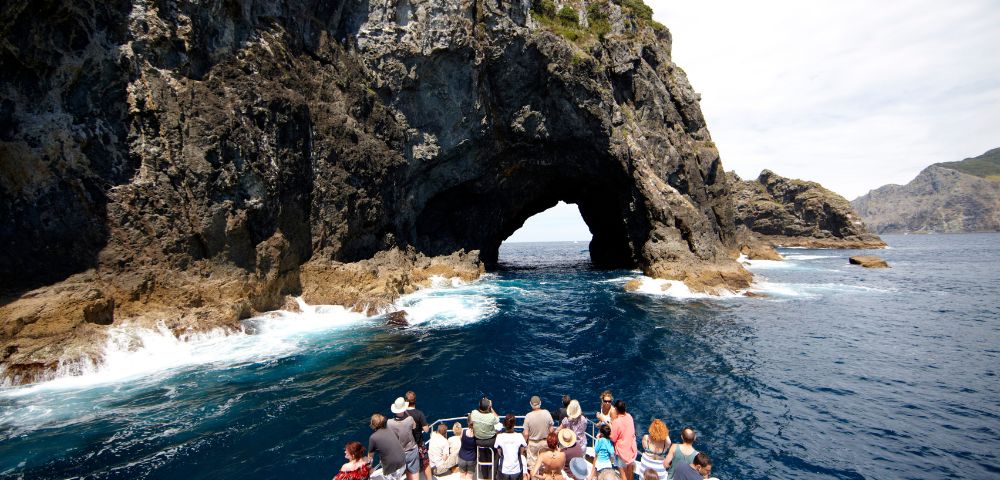 Tourists on a boat approach a large natural archway carved into a rocky cliff face over the ocean.