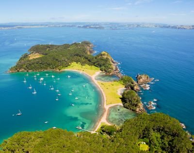 Aerial view of a lush, green island with a crescent-shaped beach and clear turquoise waters dotted with sailboats.