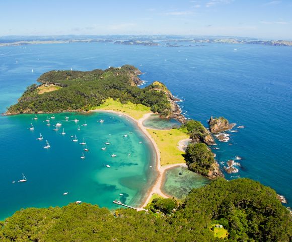 Aerial view of a lush, green island with a crescent-shaped beach and clear turquoise waters dotted with sailboats.