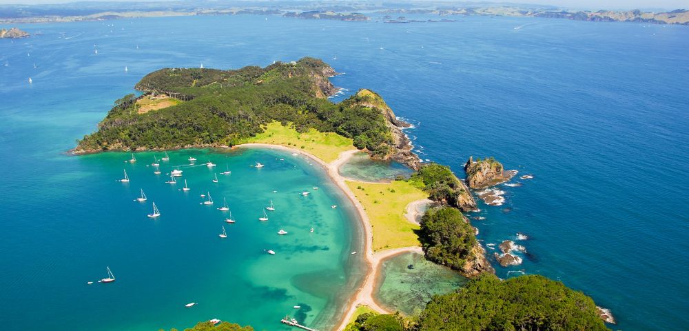 Aerial view of a lush, green island with a crescent-shaped beach and clear turquoise waters dotted with sailboats.
