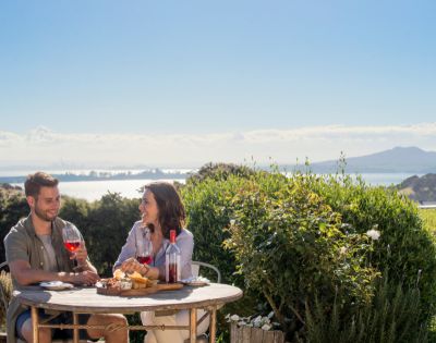 A couple enjoys wine and cheese on a patio overlooking a bay, with lush greenery in the foreground and distant mountains under a clear blue sky.