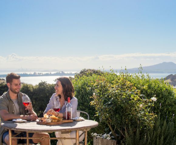 A couple enjoys wine and cheese on a patio overlooking a bay, with lush greenery in the foreground and distant mountains under a clear blue sky.