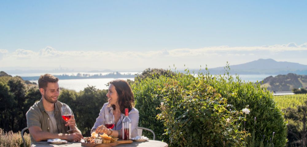 A couple enjoys wine and cheese on a patio overlooking a bay, with lush greenery in the foreground and distant mountains under a clear blue sky.