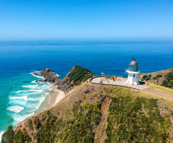Aerial view of a white lighthouse on a grassy cliff overlooking a bright blue ocean with white waves crashing on a sandy beach.