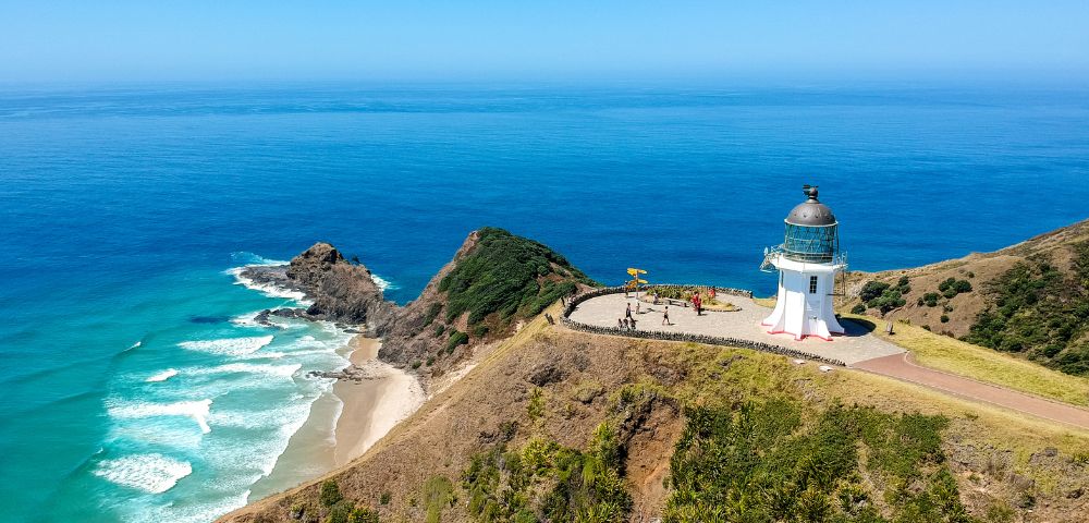 Aerial view of a white lighthouse on a grassy cliff overlooking a bright blue ocean with white waves crashing on a sandy beach.