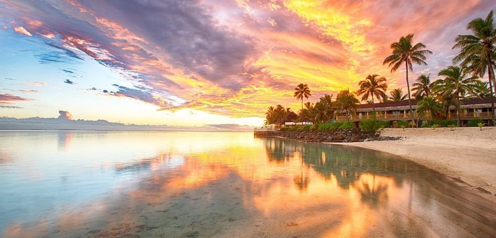 A vibrant sunset casts fiery oranges and yellows across a cloudy sky, reflected in the calm, clear ocean waters. Palm trees and a low-rise resort line a sandy beach in the foreground.