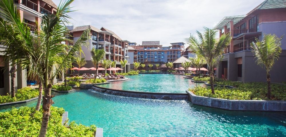 A sprawling resort complex with modern architecture surrounds a large, turquoise swimming pool. Palm trees and lush greenery are interspersed throughout the grounds, and the sky is a bright blue with scattered clouds.