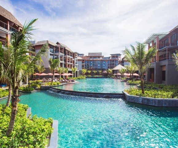 A sprawling resort complex with modern architecture surrounds a large, turquoise swimming pool. Palm trees and lush greenery are interspersed throughout the grounds, and the sky is a bright blue with scattered clouds.