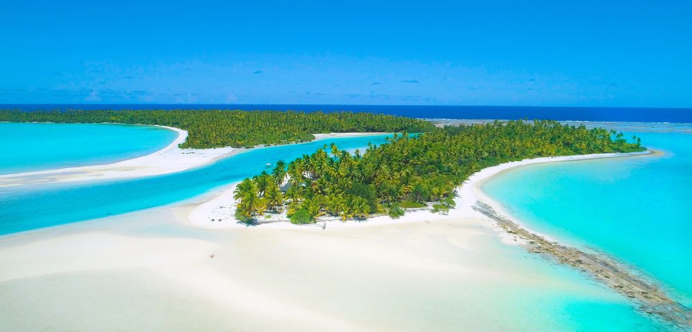 Aerial view of a tropical island with lush green palm trees, white sandy beaches, and crystal-clear turquoise lagoons under a bright blue sky.