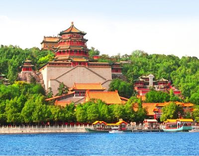 A panoramic view of a large, multi-tiered Chinese palace complex with red and gold roofs, set against a lush green hillside and a bright blue sky, with a calm blue lake in the foreground dotted with traditional boats.