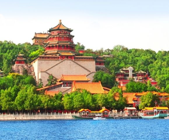 A panoramic view of a large, multi-tiered Chinese palace complex with red and gold roofs, set against a lush green hillside and a bright blue sky, with a calm blue lake in the foreground dotted with traditional boats.