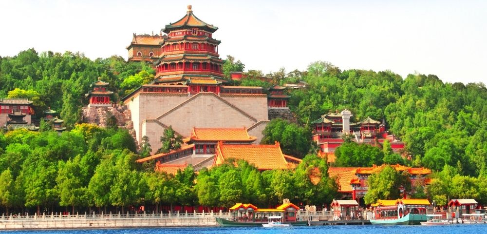 A panoramic view of a large, multi-tiered Chinese palace complex with red and gold roofs, set against a lush green hillside and a bright blue sky, with a calm blue lake in the foreground dotted with traditional boats.
