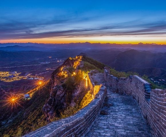 A section of the Great Wall of China illuminated by lights at dusk, winding along a ridge with a city visible in the valley below.