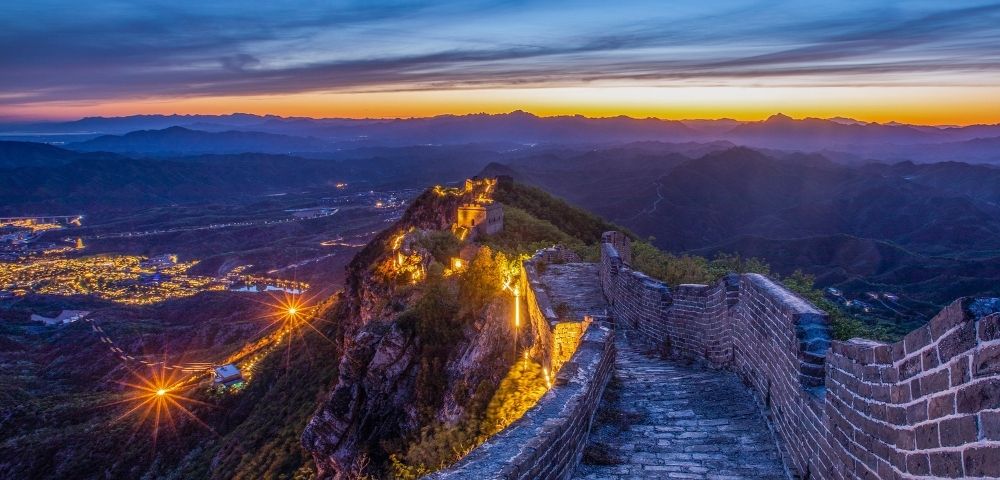 A section of the Great Wall of China illuminated by lights at dusk, winding along a ridge with a city visible in the valley below.
