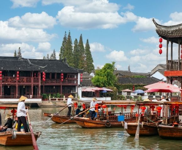 Traditional wooden boats with people in conical hats navigate a calm canal alongside ornate Chinese architecture decorated with red lanterns.