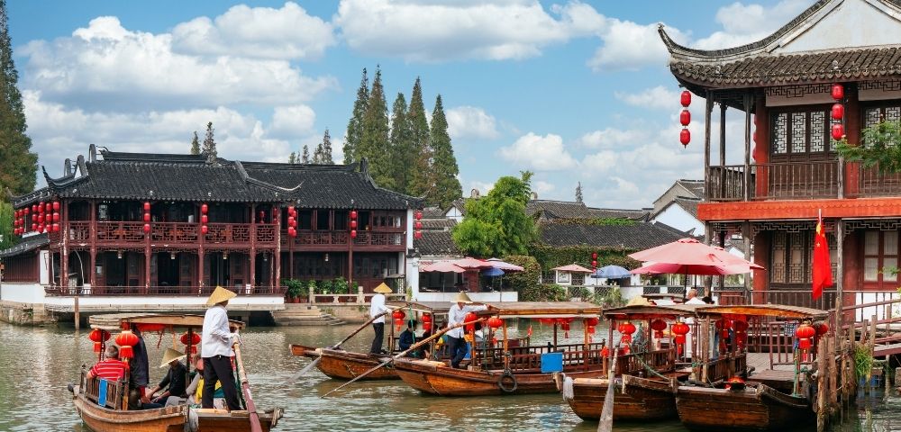 Traditional wooden boats with people in conical hats navigate a calm canal alongside ornate Chinese architecture decorated with red lanterns.