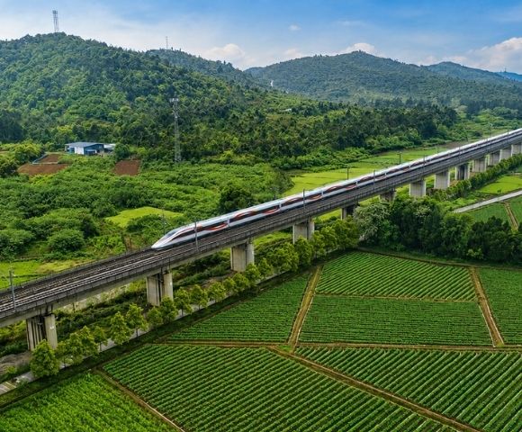 An aerial view captures a white and red high-speed train traveling on an elevated railway track, which runs through lush green agricultural fields and rolling forested hills under a blue sky with scattered clouds.