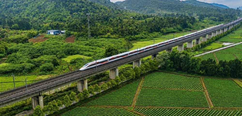 An aerial view captures a white and red high-speed train traveling on an elevated railway track, which runs through lush green agricultural fields and rolling forested hills under a blue sky with scattered clouds.