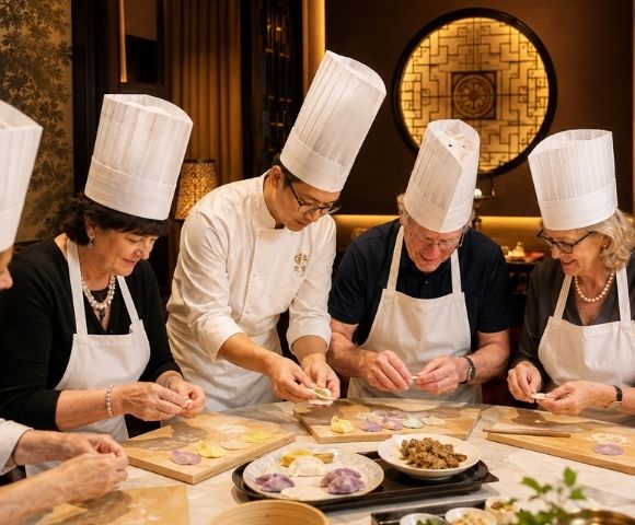A chef in a white uniform and toque instructs several adults in white aprons and toques as they prepare colorful dumplings on wooden boards in a well-lit, elegant dining room.