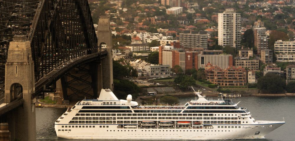 A large white cruise ship sails on a body of water with the Sydney Harbour Bridge and cityscape in the background.