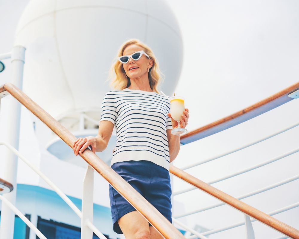 Woman with blonde hair and white sunglasses holding a yellow drink, standing on the stairs of a cruise ship.