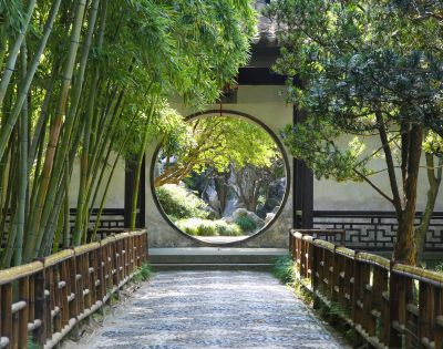 A circular opening in a wall frames a view of a serene garden with trees and rocks, with a pathway leading towards it, bordered by bamboo on the left and a wooden fence on both sides.
