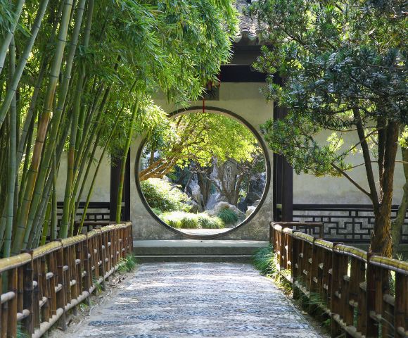 A circular opening in a wall frames a view of a serene garden with trees and rocks, with a pathway leading towards it, bordered by bamboo on the left and a wooden fence on both sides.