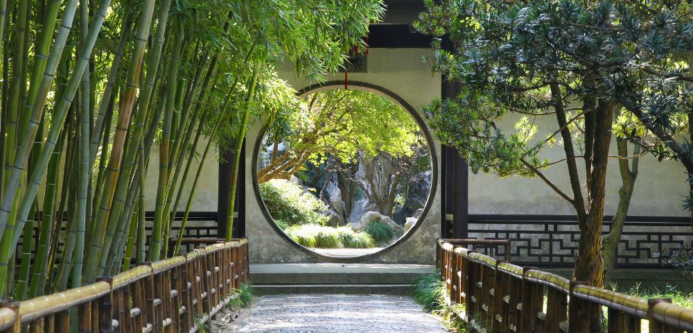 A circular opening in a wall frames a view of a serene garden with trees and rocks, with a pathway leading towards it, bordered by bamboo on the left and a wooden fence on both sides.