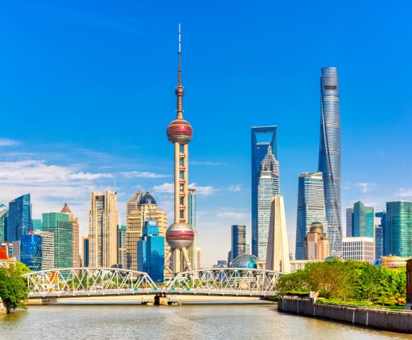 Panoramic view of the Shanghai skyline with the Oriental Pearl TV Tower and other modern skyscrapers under a bright blue sky, with a bridge spanning a river in the foreground.