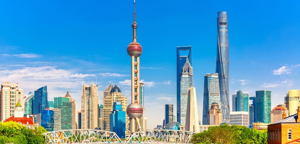 Panoramic view of the Shanghai skyline with the Oriental Pearl TV Tower and other modern skyscrapers under a bright blue sky, with a bridge spanning a river in the foreground.