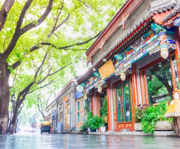 Traditional Chinese buildings with ornate colorful eaves line a wet cobblestone street shaded by large trees with lush green leaves.