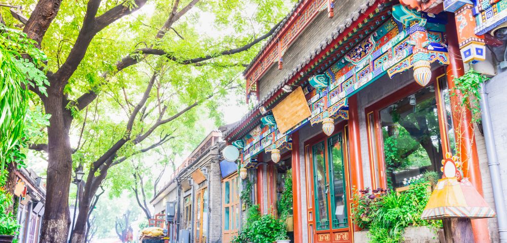 Traditional Chinese buildings with ornate colorful eaves line a wet cobblestone street shaded by large trees with lush green leaves.