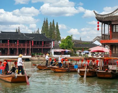 Wooden boats with people wearing conical hats glide along a canal beside traditional East Asian architecture with red lanterns and tiled roofs under a cloudy sky.