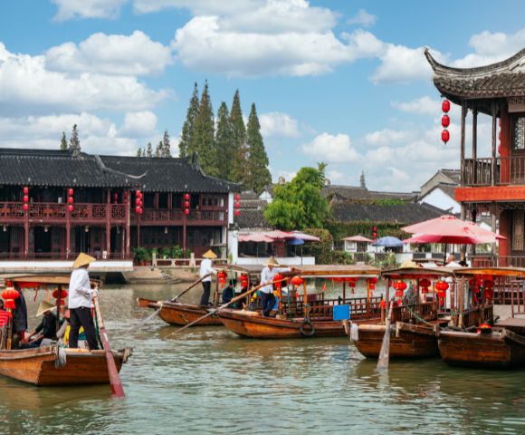 Wooden boats with people wearing conical hats glide along a canal beside traditional East Asian architecture with red lanterns and tiled roofs under a cloudy sky.