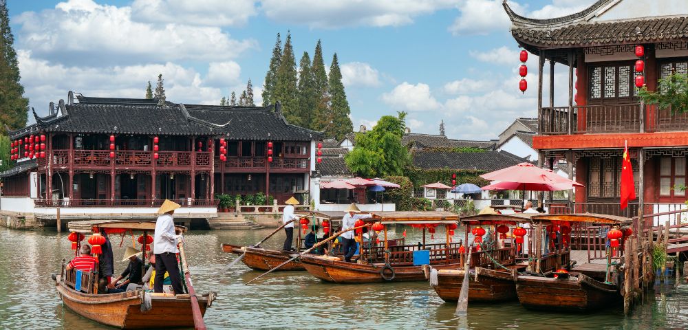 Wooden boats with people wearing conical hats glide along a canal beside traditional East Asian architecture with red lanterns and tiled roofs under a cloudy sky.