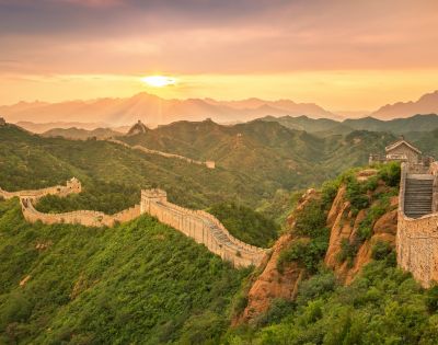 The Great Wall of China winds across lush green, mountainous terrain under a hazy sunset sky.
