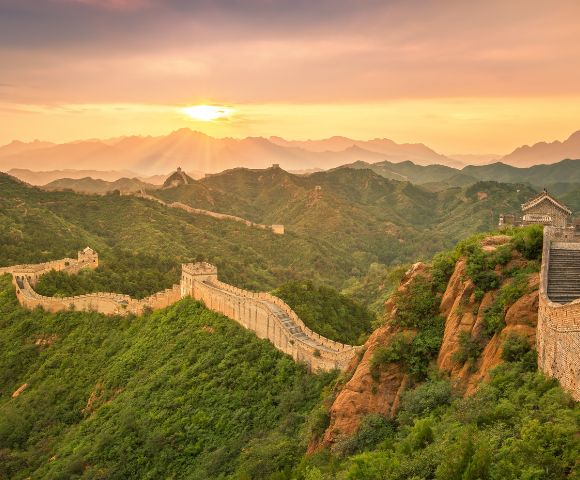 The Great Wall of China winds across lush green, mountainous terrain under a hazy sunset sky.