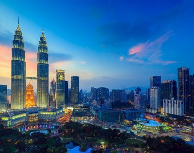 Panoramic view of a modern city skyline at dusk, featuring the illuminated Petronas Twin Towers, surrounded by numerous skyscrapers and a park with lush trees and water features, under a vibrant blue and pink sky.